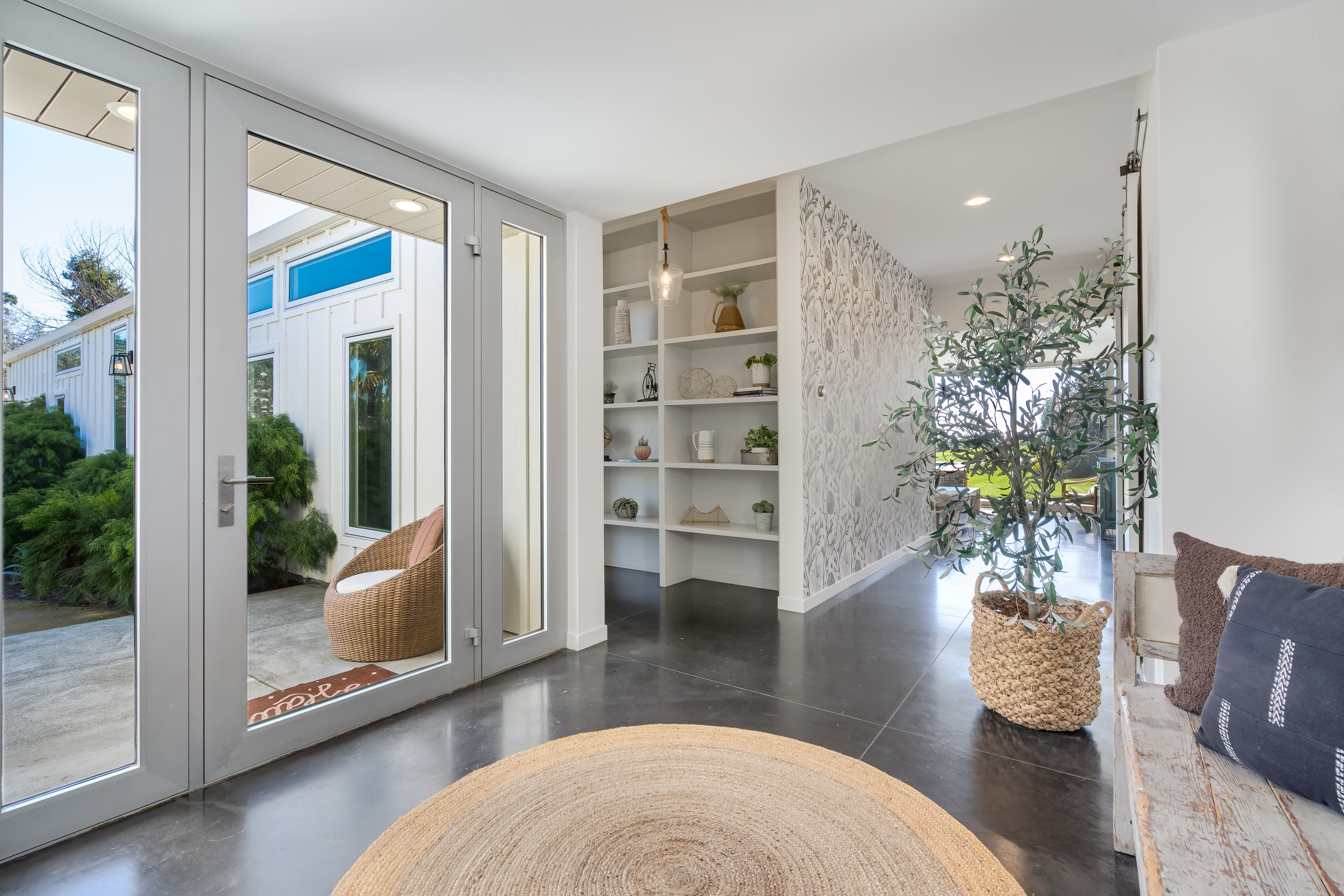 Sunlit modern entryway with glass double doors to a patio, round jute rug on polished concrete, wicker patio chair outside, built-in white shelving, patterned accent wall, woven-basket potted plant and rustic bench with pillows.
