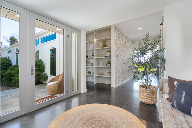 Sunlit modern entryway with glass double doors to a patio, round jute rug on polished concrete, wicker patio chair outside, built-in white shelving, patterned accent wall, woven-basket potted plant and rustic bench with pillows.