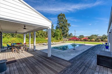 Sunny modern suburban backyard with rectangular swimming pool, concrete deck and red umbrellas, wooden wraparound deck and covered dining porch with ceiling fans and outdoor table
