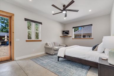 Bright modern bedroom with dark wood bed and white linens on a blue area rug, slipcovered armchair, ceiling fan, wall-mounted TV, wood-trim windows and glass door opening to a backyard patio.