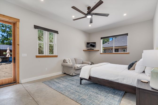 Bright modern bedroom with dark wood bed and white linens on a blue area rug, slipcovered armchair, ceiling fan, wall-mounted TV, wood-trim windows and glass door opening to a backyard patio.