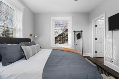 Sunlit modern bedroom with a gray-upholstered bed dressed in gray and white bedding and striped lumbar pillow, brass wall sconce, window with white blinds showing nearby houses, leaning full-length mirror, wall-mounted TV and dark hardwood floors.