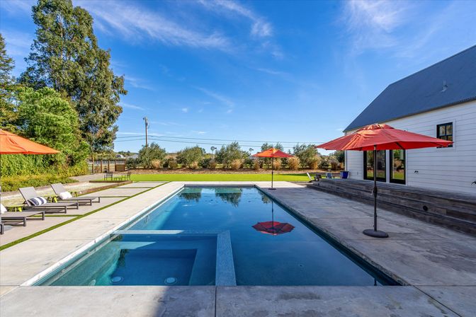Sunlit suburban backyard pool with built-in spa, concrete deck, red patio umbrellas, lounge chairs and manicured lawn under a clear blue sky