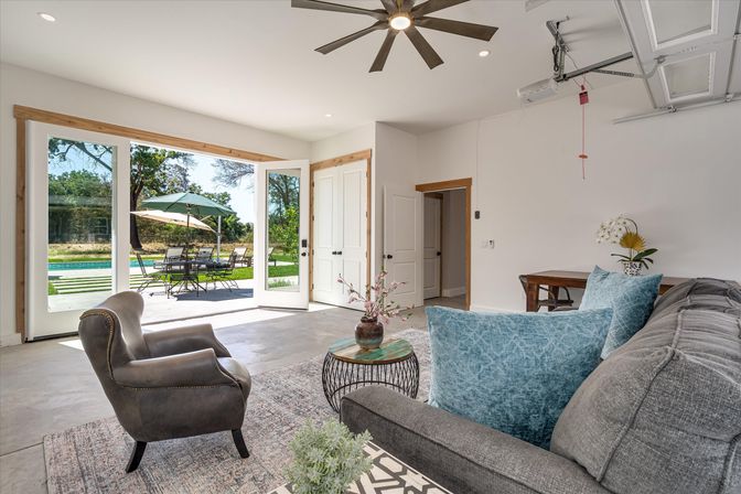 Bright modern open-plan living room with large glass doors opening to a sunny backyard patio and pool, gray sofa with blue pillows, leather armchair, round wire coffee table, ceiling fan and outdoor dining umbrella