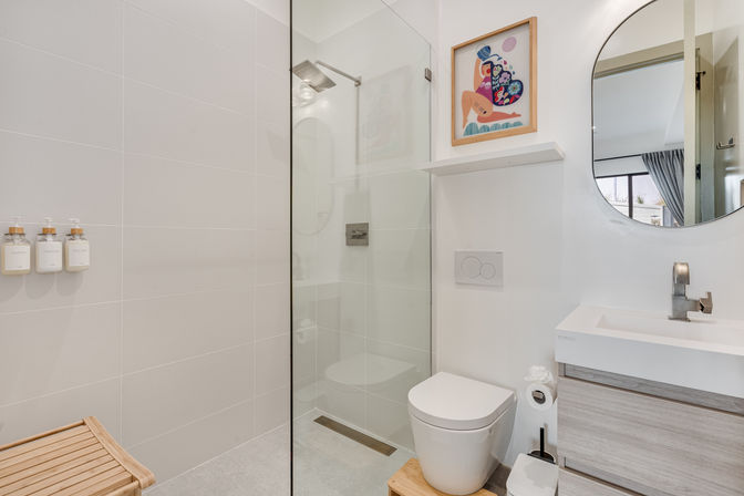 Bright modern minimalist bathroom with glass walk-in shower and rainhead, wall-mounted dispensers, white toilet, wood-tone vanity, round mirror and colorful wall art, cheerful vibe.