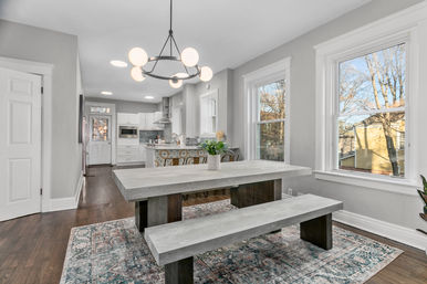 Bright open-concept dining area in a suburban home with a concrete-style farmhouse table and bench on a patterned rug, globe chandelier, hardwood floors, large white-trim windows overlooking trees, and a white kitchen with patterned tile island and stainless appliances in the background.