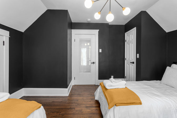 Cozy modern attic bedroom with dark charcoal walls, white trim and hardwood floors, twin beds dressed in white linens with mustard-yellow throws, mid-century chandelier and glass-paneled door.