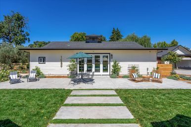 Sunny suburban backyard patio of a modern single-story home with white siding and glass French doors, a concrete stepping-stone path across a green lawn, outdoor dining set with umbrella and wicker lounge chairs.