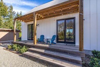 Sunlit modern farmhouse porch with wooden pergola and string lights, blue Adirondack chairs on a weathered wood deck, black sliding glass doors and white board-and-batten siding overlooking a gravel yard.