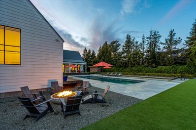 Cozy modern farmhouse backyard at dusk with a lit circular fire pit surrounded by Adirondack chairs, rectangular swimming pool with red umbrellas, concrete patio and manicured lawn framed by tall trees.