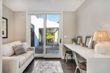 Bright, cozy living room and home office nook with beige sofa and white desk, sliding glass doors opening to a covered wooden deck with outdoor dining table and green yard