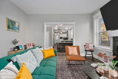 Open-plan modern living room with a teal tufted sofa and mustard throw pillows, tan leather accent chair, round coffee table with plants, hardwood floors and large window, looking into a contemporary kitchen with island and statement pendant light.
