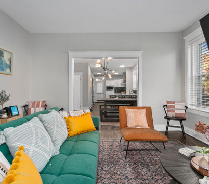 Open-plan modern living room with a teal tufted sofa and mustard throw pillows, tan leather accent chair, round coffee table with plants, hardwood floors and large window, looking into a contemporary kitchen with island and statement pendant light.