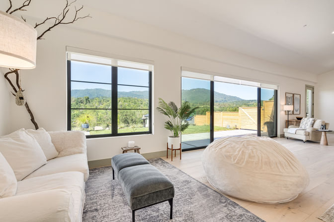 Bright modern living room with white sofa, oversized cream beanbag and gray ottoman, black-framed windows and sliding glass doors opening to a sunny patio, grassy yard and distant green hills.
