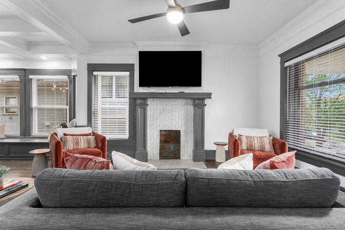 Cozy modern living room with a gray sofa facing a herringbone-tile fireplace topped by a wall TV, two rust velvet armchairs, large windows with blinds and a ceiling fan.