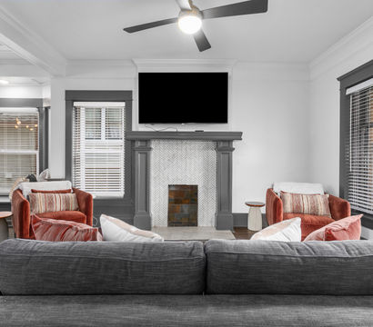 Cozy modern living room with a gray sofa facing a herringbone-tile fireplace topped by a wall TV, two rust velvet armchairs, large windows with blinds and a ceiling fan.