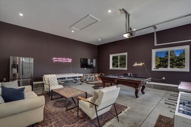 Spacious garage-style game room with pool table, air hockey table, foosball, comfy lounge seating, neon wall signs, concrete floor and windows overlooking trees.