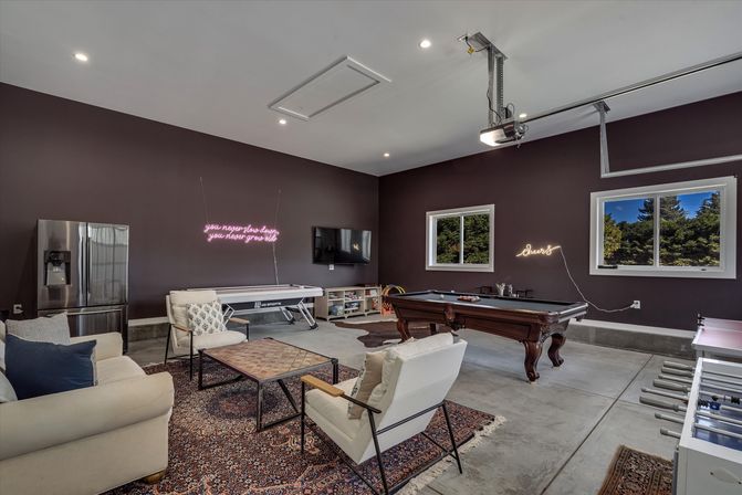 Spacious garage-style game room with pool table, air hockey table, foosball, comfy lounge seating, neon wall signs, concrete floor and windows overlooking trees.