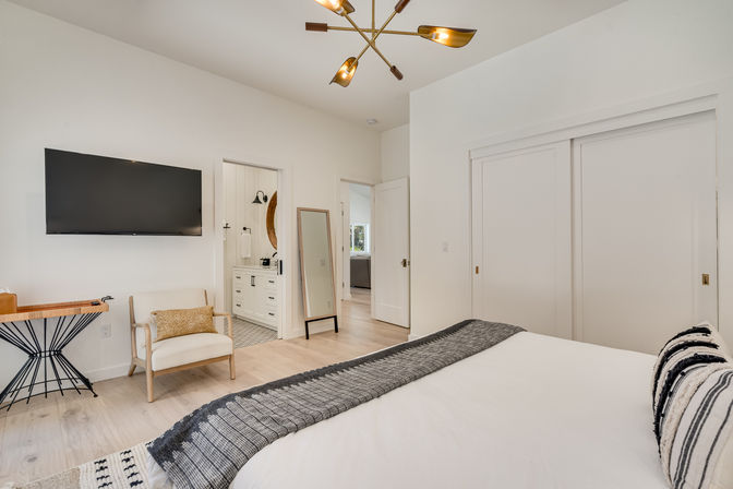 Bright modern bedroom with neutral decor, wall-mounted TV, cozy armchair with accent pillow, full-length mirror, sliding closet doors, light wood floors, brass chandelier, and doorway to en-suite bathroom.