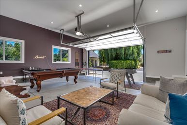 Bright garage game room with wooden pool table, foosball and ping-pong tables, comfy lounge seating on a patterned rug, neon "cheers" wall sign, and a large open garage door overlooking a landscaped yard with a willow tree.