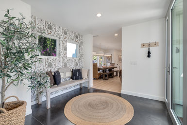 Bright modern home entryway/foyer with botanical wallpaper, weathered wooden bench with throw pillows, round jute rug, woven basket and potted olive tree, flooded with natural light and a view into a farmhouse-style dining room.
