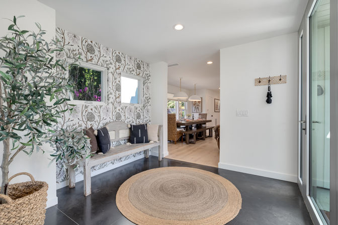 Bright modern home entryway/foyer with botanical wallpaper, weathered wooden bench with throw pillows, round jute rug, woven basket and potted olive tree, flooded with natural light and a view into a farmhouse-style dining room.