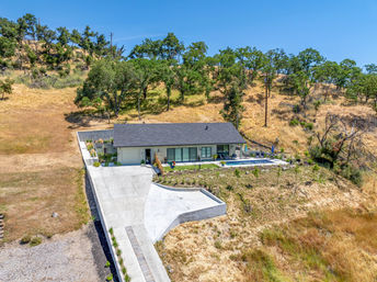 Aerial view of a modern single-story ranch home with pool and long concrete driveway on a sunny oak-studded dry hillside.