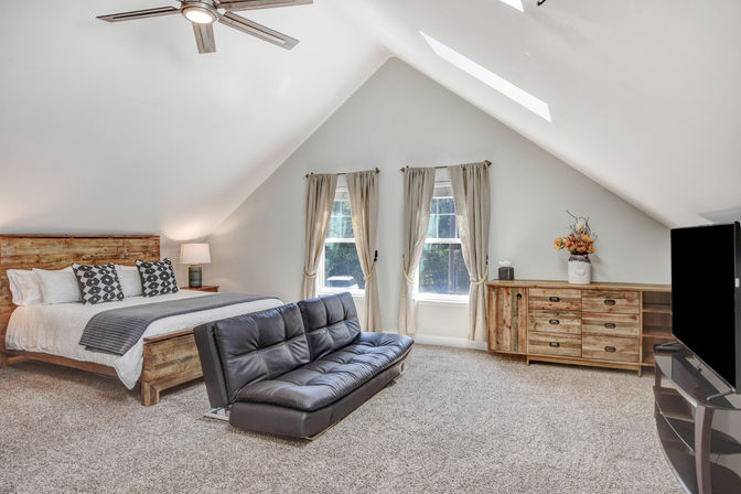 Cozy bright attic master bedroom with vaulted ceiling and skylights, rustic wooden bed and dresser, black leather convertible sofa, carpeted floor, two tall windows with beige curtains, ceiling fan and flat-screen TV.