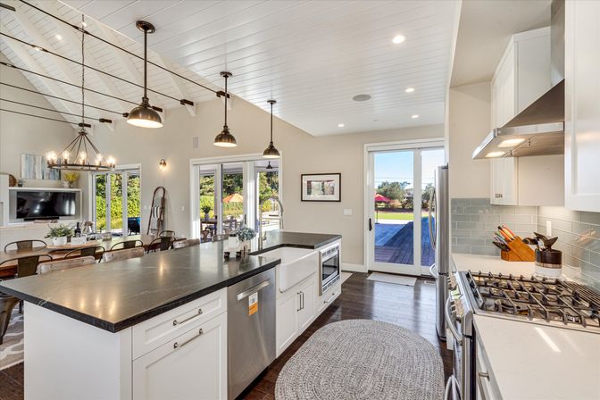 Open-concept modern farmhouse kitchen with large dark island, farmhouse sink, stainless appliances, pendant lights, dining table and sliding glass doors to backyard patio and pool.