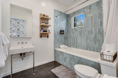 Modern spa-like bathroom with white sink, bathtub surrounded by blue-green vertical subway tiles, small window, wooden wall shelves with toiletries, white toilet, rolled towels and gray bath mat.