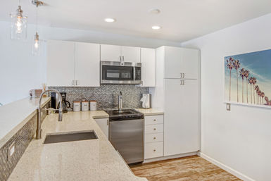 Bright modern white kitchen with quartz countertops, mosaic tile backsplash, stainless steel microwave and dishwasher, undermount sink with gooseneck faucet, glass pendant lights, palm-print wall art and wood-look flooring.