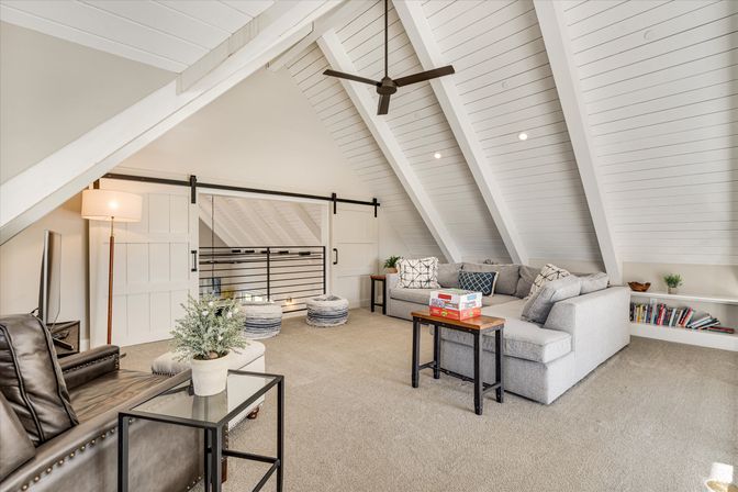 Bright cozy attic loft living area with white vaulted shiplap ceiling and exposed beams, gray L-shaped sectional, barn-style sliding door, ceiling fan, carpeted floor, side table with board games and low bookshelf.