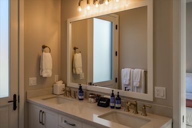 Cozy residential double-sink bathroom with marble countertop, brass faucets, large framed mirror, frosted glass door, warm globe vanity lights and neatly folded white towels.