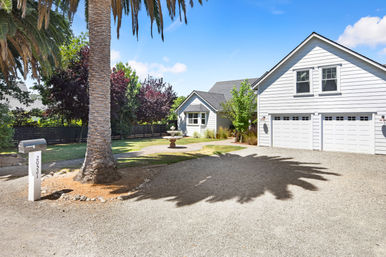 Sunny suburban white two-story house with three-car garage and gravel driveway, large palm tree casting a bold shadow, front-yard fountain, mailbox, and leafy trees under a blue sky.
