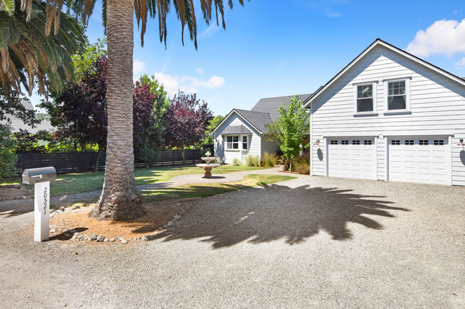 Sunny suburban white two-story house with three-car garage and gravel driveway, large palm tree casting a bold shadow, front-yard fountain, mailbox, and leafy trees under a blue sky.
