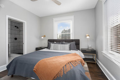 Bright, cozy contemporary gray bedroom with dark upholstered headboard, blue-gray bedding and orange throw blanket, brass wall sconces, matching nightstands, hardwood floors, window with white blinds, and an open doorway to a tiled en-suite shower.