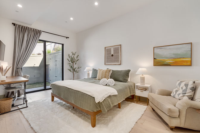 Bright, spa-like contemporary bedroom in a modern apartment — sage-green bed with rolled towels, sliding glass door to a small patio, desk workspace, comfy armchair, abstract wall art, and plush white rug.