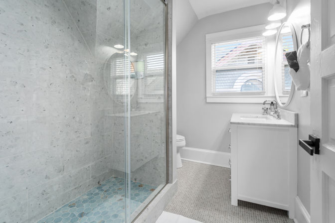 Bright modern residential bathroom with glass walk-in shower, marble-look wall tile, blue hexagon shower floor, white vanity with round mirror, window blinds, and patterned floor tiles