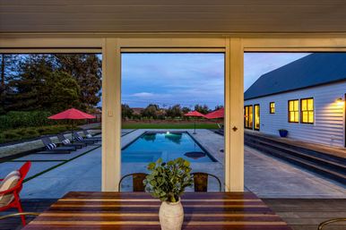 Dusk view from a covered patio over a wooden dining table with a potted plant, looking out to a rectangular backyard pool, lounge chairs with red umbrellas and a white house with glowing windows.