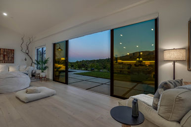Cozy open-plan modern bedroom-living area with floor-to-ceiling sliding glass doors opening to a patio and grassy yard at sunset, neutral furnishings, potted plants, soft lamp lighting and light wood floors.