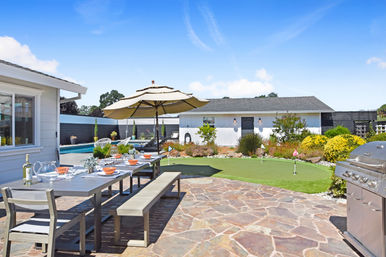 Sunny backyard patio with stone pavers, long outdoor dining table set for al fresco meals under a large umbrella, stainless-steel grill, swimming pool, small putting green and landscaped shrubs in front of a white pool house.
