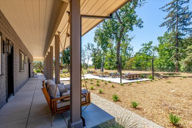 Inviting sunlit covered porch with a cushioned wooden swing on a columned walkway overlooking a landscaped yard and a concrete outdoor lounge area with benches and tall trees