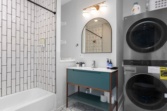 Bright modern bathroom with stacked washer-dryer, bathtub-shower with vertical white subway tile, teal vanity with arched mirror, and geometric floor tile.