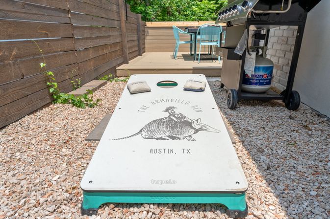 Outdoor cornhole board featuring an armadillo illustration and “Austin, TX” lettering on a pebbled yard, beside a wooden fence, a deck with teal chairs, and a propane grill.