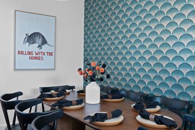 Modern dining room with teal scallop-pattern wallpaper, wooden table set with plates and black napkins, white vase of orange flowers, and a playful framed armadillo poster reading "ROLLING WITH THE HOMIES".