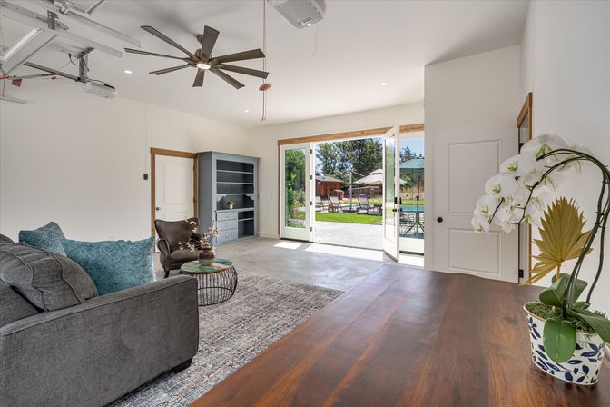 Bright open-plan living room with gray sofa, teal pillows, leather armchair and round coffee table, large ceiling fan and shelving, folding glass doors opening to a sunny backyard patio with lounge chairs and umbrellas, white orchid on a wooden counter in the foreground.