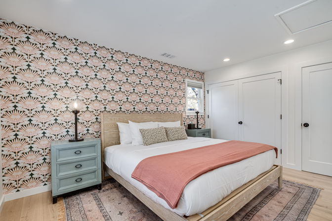 Stylish bedroom with pink-and-green floral wallpaper accent wall, light wood platform bed dressed in white linens and a coral throw, mint bedside tables with vintage lamps, patterned area rug and white closet doors.