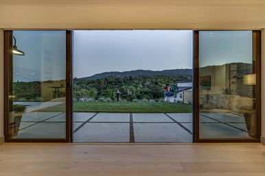 View through wide sliding glass doors to a modern patio with square concrete pavers, small lawn, birdbath and colorful yard sculpture, framed by wooded hills and a dusky mountain ridge beyond.
