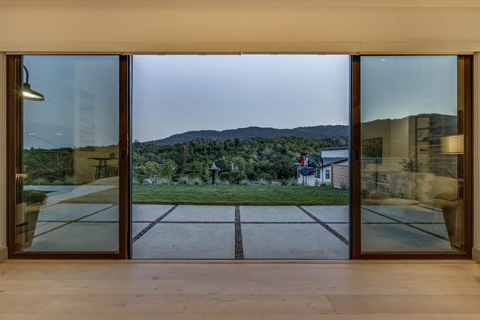 View through wide sliding glass doors to a modern patio with square concrete pavers, small lawn, birdbath and colorful yard sculpture, framed by wooded hills and a dusky mountain ridge beyond.