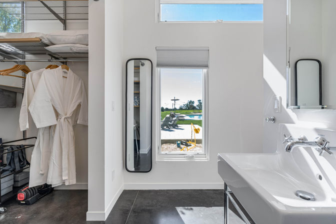 Modern bathroom and walk-in closet with a white robe on a hanger, slim full-length mirror, white sink, and a narrow window framing a sunny patio with lounge chairs, pool, and yellow sand toys.